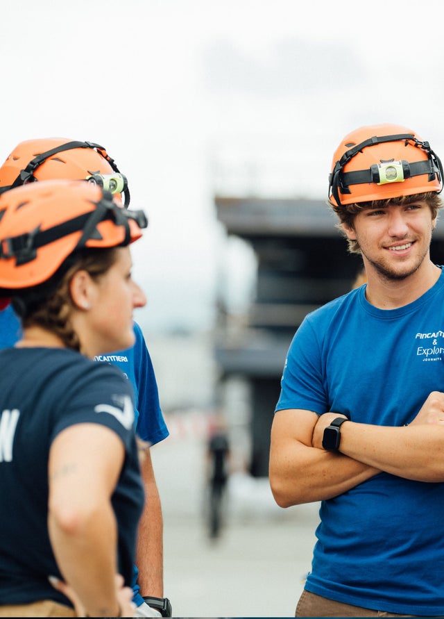 Fincantieri | Group - Sustainability - Industrial Excellence - Health and Safety: Group of people wearing orange safety helmets and blue shirts, gathered in a construction site or outdoor industrial location with scaffolding in the background, one person with a smartwatch, the shirts display the text "Pinnacle... Explore". Team atmosphere and professional readiness.