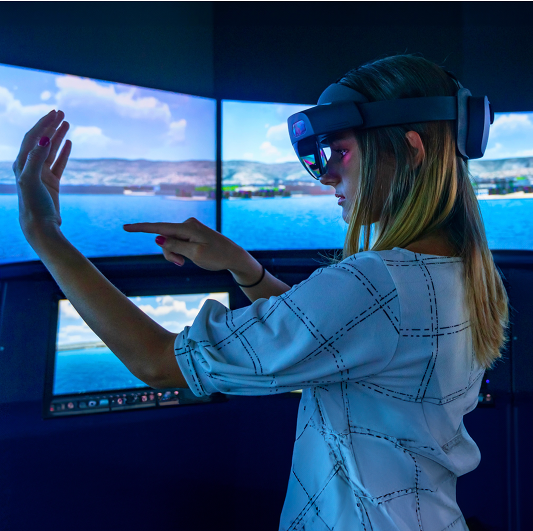 Fincantieri | Group - Innovation - Smart Manufacturing System - Robotics, Drones and Automation: Woman wearing a VR headset interacts in a high-tech simulation room, multiple screens display a coastal landscape with water and hills. Immersive gestures, bright environment, and advanced technology.