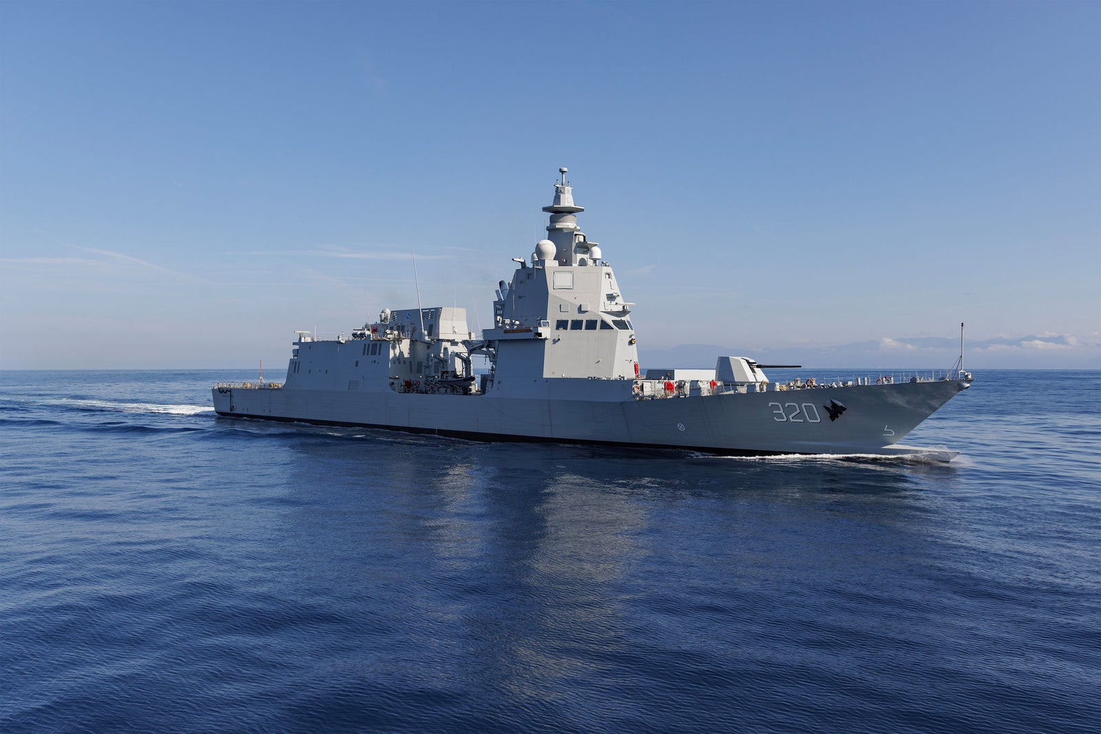 Fincantieri | Group - Companies - Subsidiaries and Affiliates - Fincantieri Naval Services: Large operational military ship '320' sailing on calm waters under a clear blue sky, modern military design and advanced equipment, maritime environment highlighting readiness and purpose.