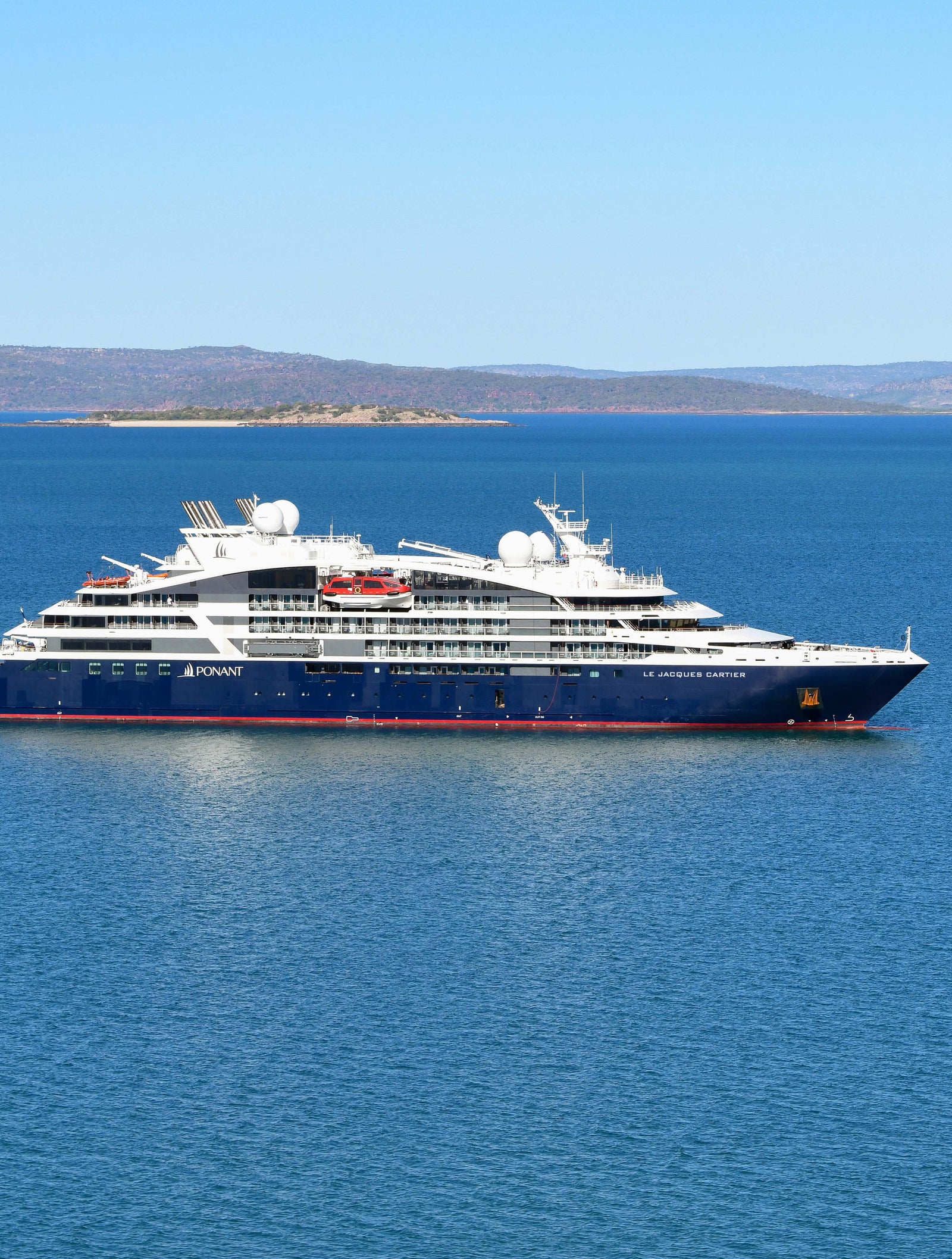  Fincantieri | Group - Business - Products - Cruise Ships - Le Jacques Cartier: Sleek luxury cruise ship in operation floating on calm clear blue waters under bright sky, modern design with multiple decks and white accents, distant landforms visible on horizon, tranquil setting.