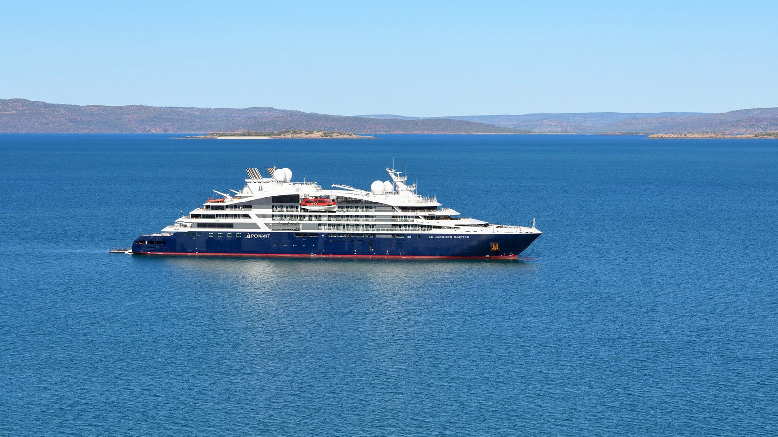 Fincantieri | Group - Business - Products - Cruise Ships - Le Jacques Cartier: View of Le Jacques Cartier cruise ship in operation, navigating calm waters, modern hull design, multiple passenger decks, mainland visible on horizon.