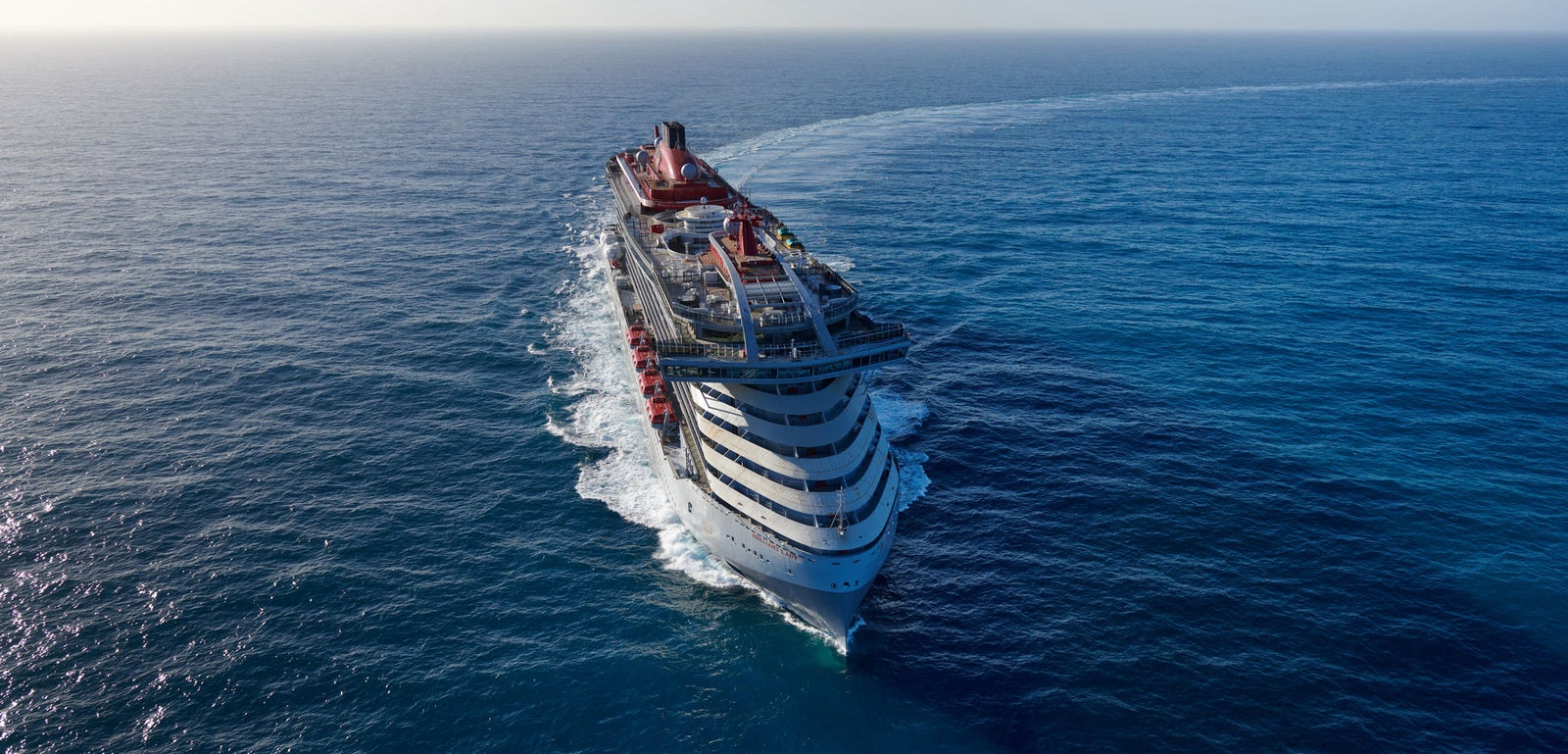 Fincantieri | Group - Business - Products - Cruise Ships - Brilliant Lady: Large modern operational cruise ship with red accents and multiple decks, sailing in calm waters under clear sky, distant mountains and wide maritime horizon.