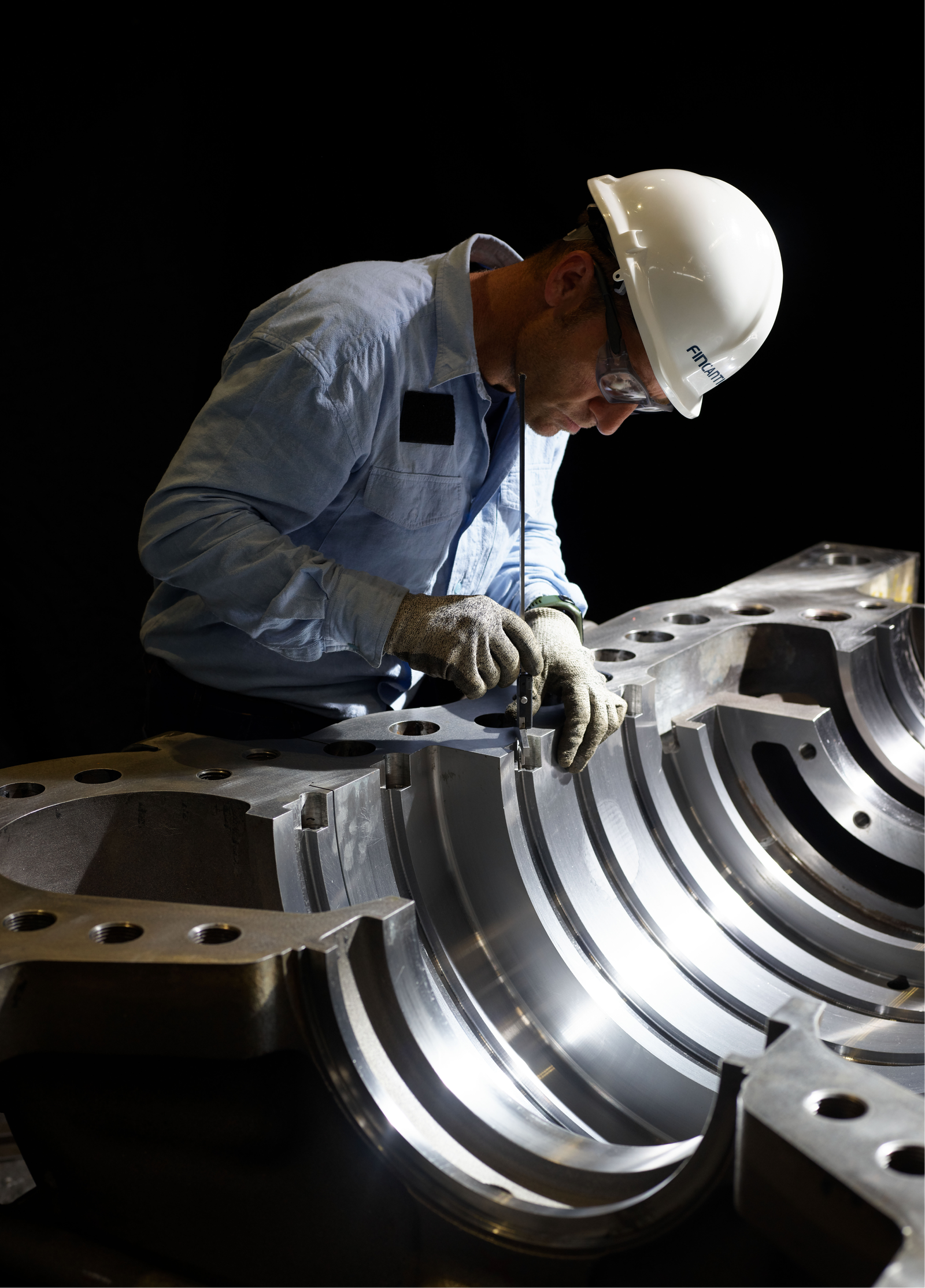 Fincantieri | Group - Sustainability - People and Community - Our People: Worker wearing protective gear observes a large metallic component with curved surfaces and visible bolts, dark industrial environment, lighting highlights precision and manufacturing quality.