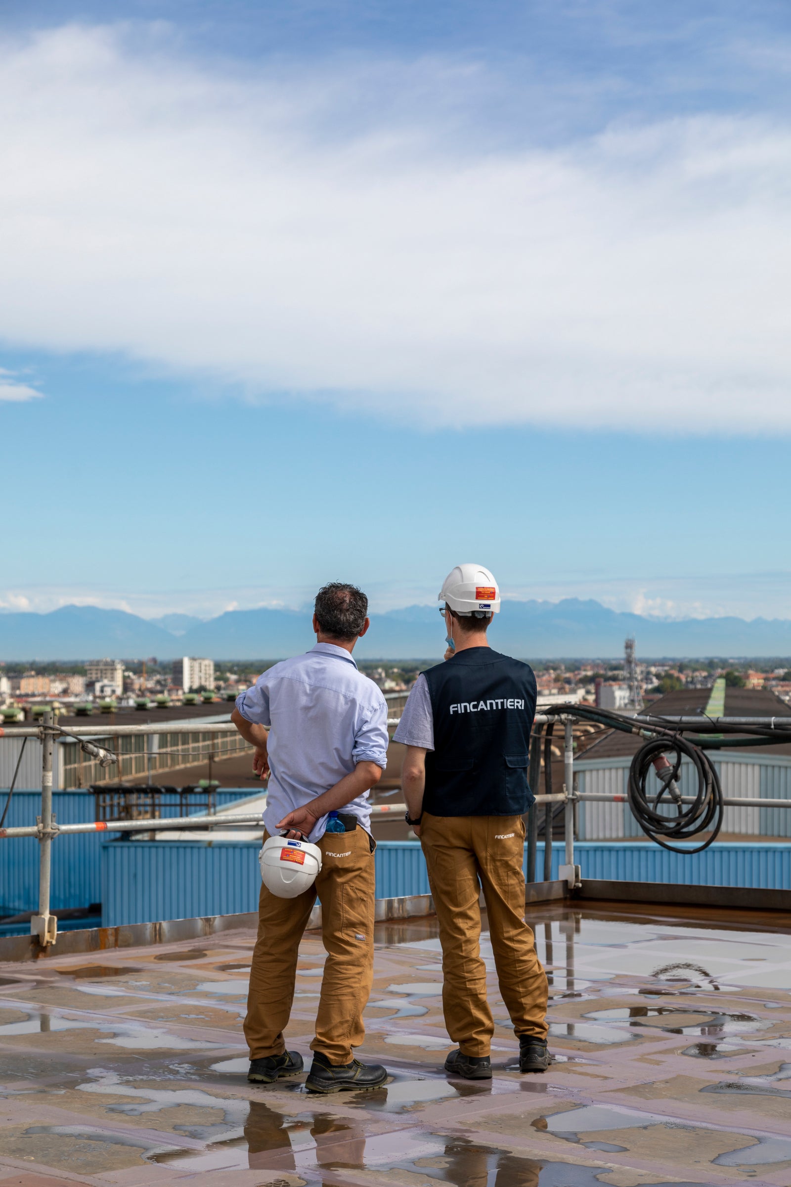 Fincantieri | People – Work With Us: Two operators wearing safety helmets on a wet platform, one with a visible “Fincantieri” vest, observing an industrial site; background with blue sky and distant mountains. Work with us