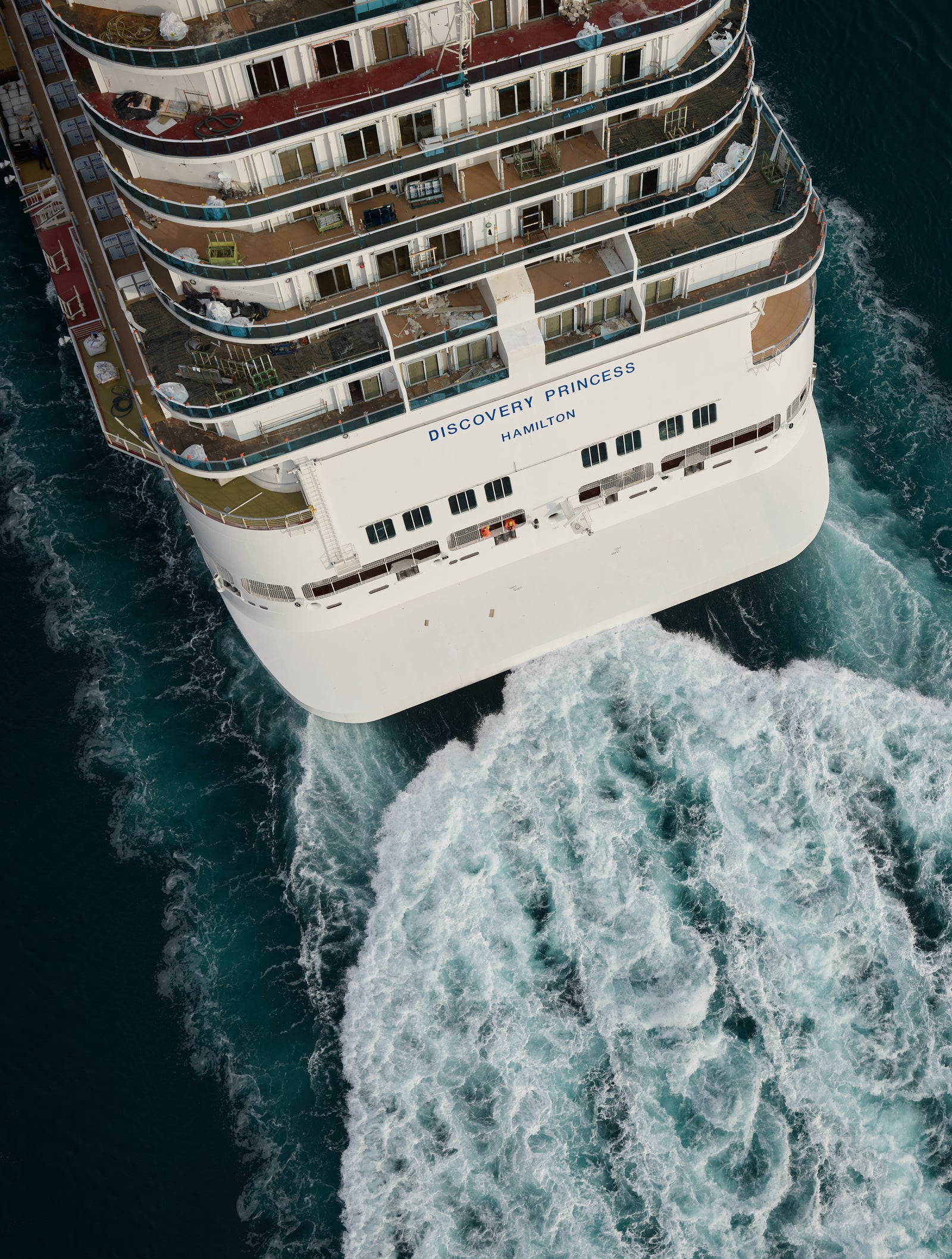 Fincantieri | Group - Business - Products - Cruise Ships - Discovery Princess: Aerial stern view of Discovery Princess cruise ship in operation, sailing on calm open waters under cloudy sky, multiple decks, red lifeboats and visible name text.