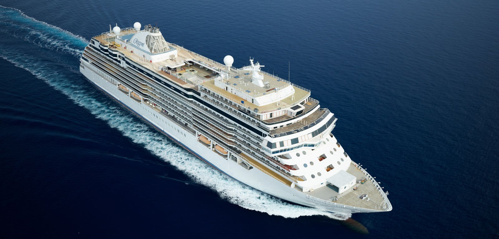 Fincantieri | Group - Business - Products - Cruise Ships - Seven Seas Splendor: Large modern operational white cruise ship with multiple decks and visible lifeboats, sailing on calm waters under clear and serene sky, visible wake, wide horizon and marine landscape.