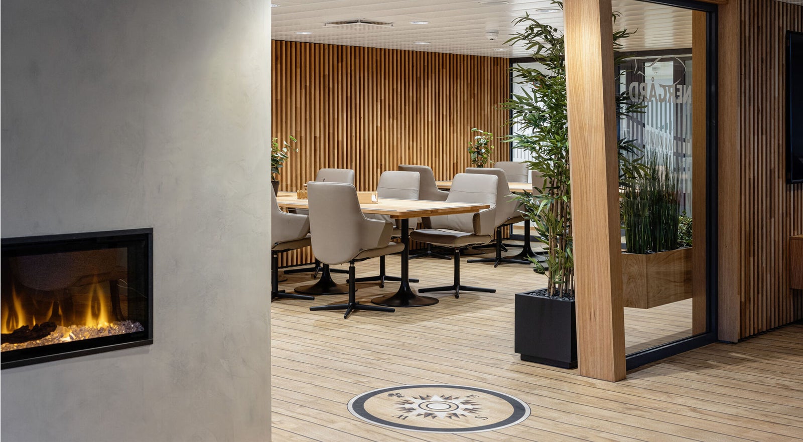 Fincantieri | Gruppo - Società - Controllate e collegate - Vard Interiors: Lounge ufficio contemporanea con parete in doghe di legno e camino elettrico, area meeting con sedie beige e tavoli in legno, presenza di piante d’interno; pavimento circolare e divisori in vetro accentuano l’estetica moderna.