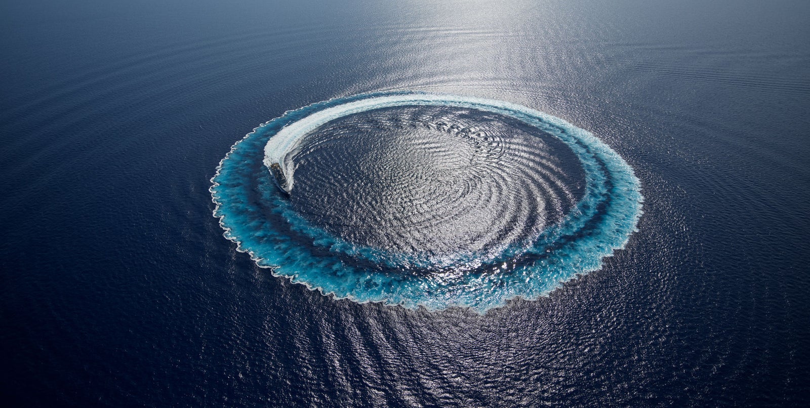 Fincantieri | About Us - Future on Board: Aerial view of a speedboat tracing a perfect circular wake on deep blue ocean, the white trail creates a distinctive visual pattern, contrast between the water and the movement of the vessel.