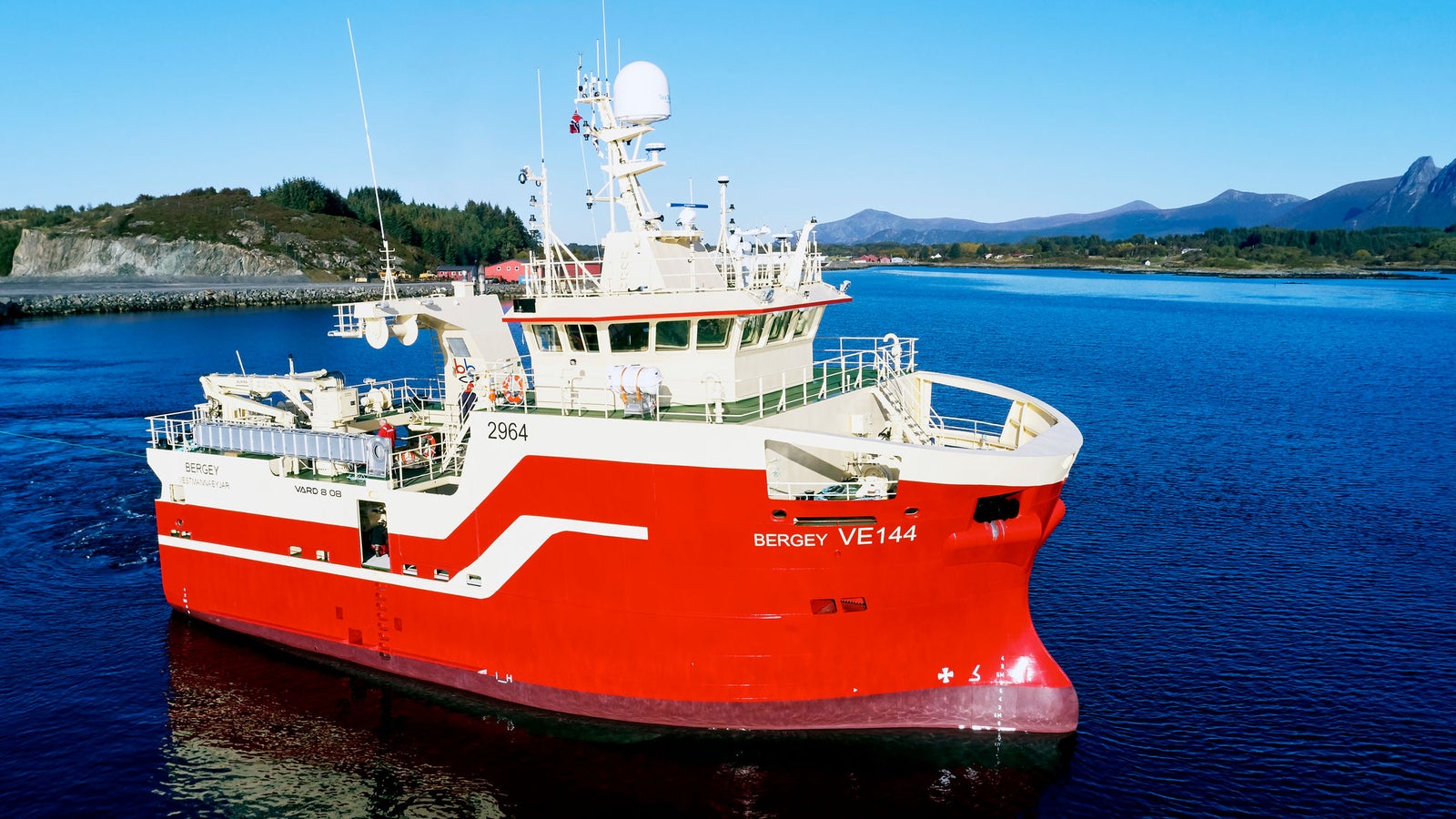 Prodotto - Peschereccio Bergey VE144: Vista laterale della nave operativa in navigazione su acque costiere, scafo rosso con banda bianca, scritte Sergey VE144 e numero 2964 visibili, sfondo verdeggiante con montagne e cielo azzurro.