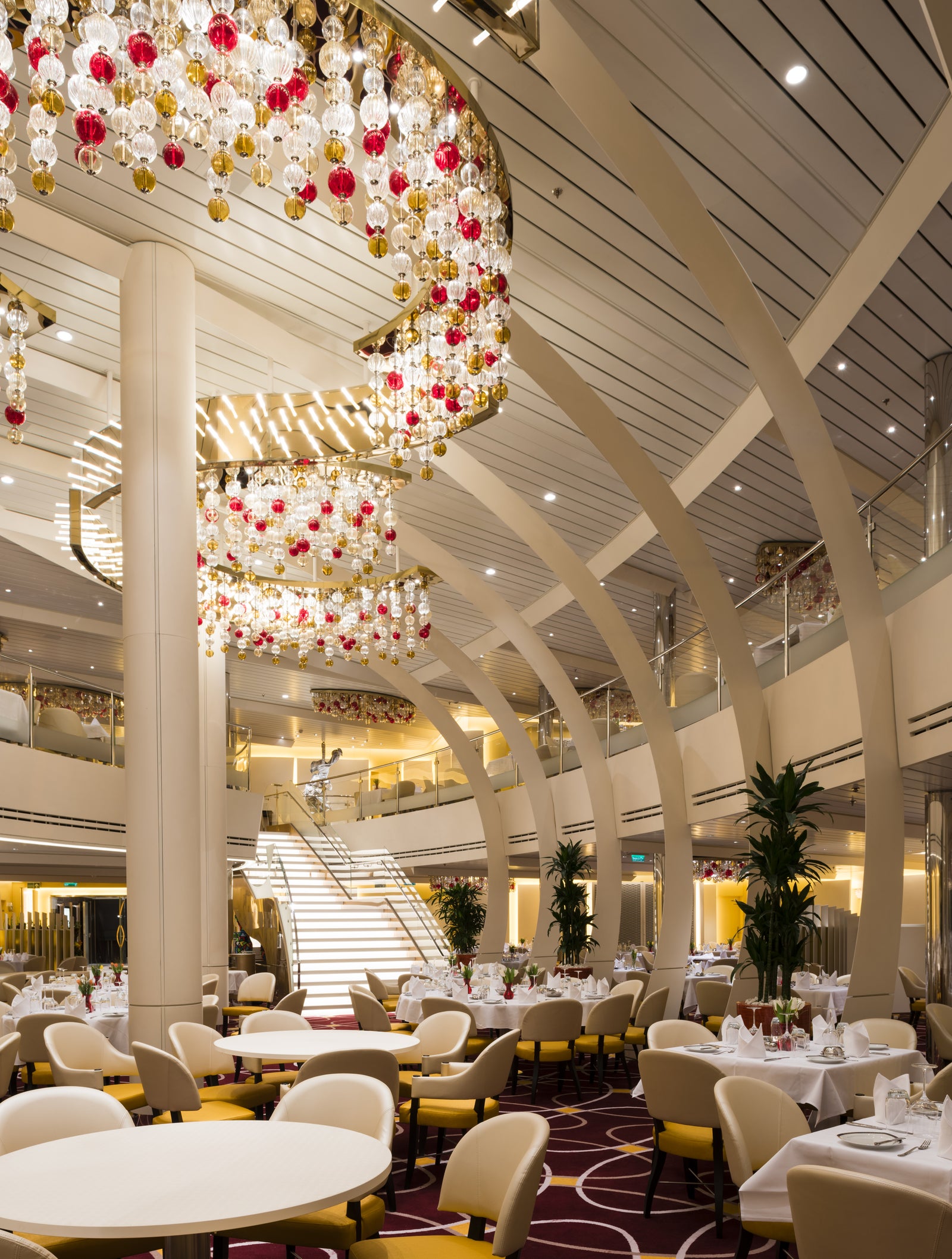 Fincantieri | Gruppo - Business - Prodotti - Navi da Crociera - Koningsdam: Spazio ristorante lussuoso con grandi lampadari circolari decorati con ornamenti in vetro rossi e trasparenti, tavoli e sedie bianchi su tappeto bordeaux decorato, scala che conduce al piano superiore.