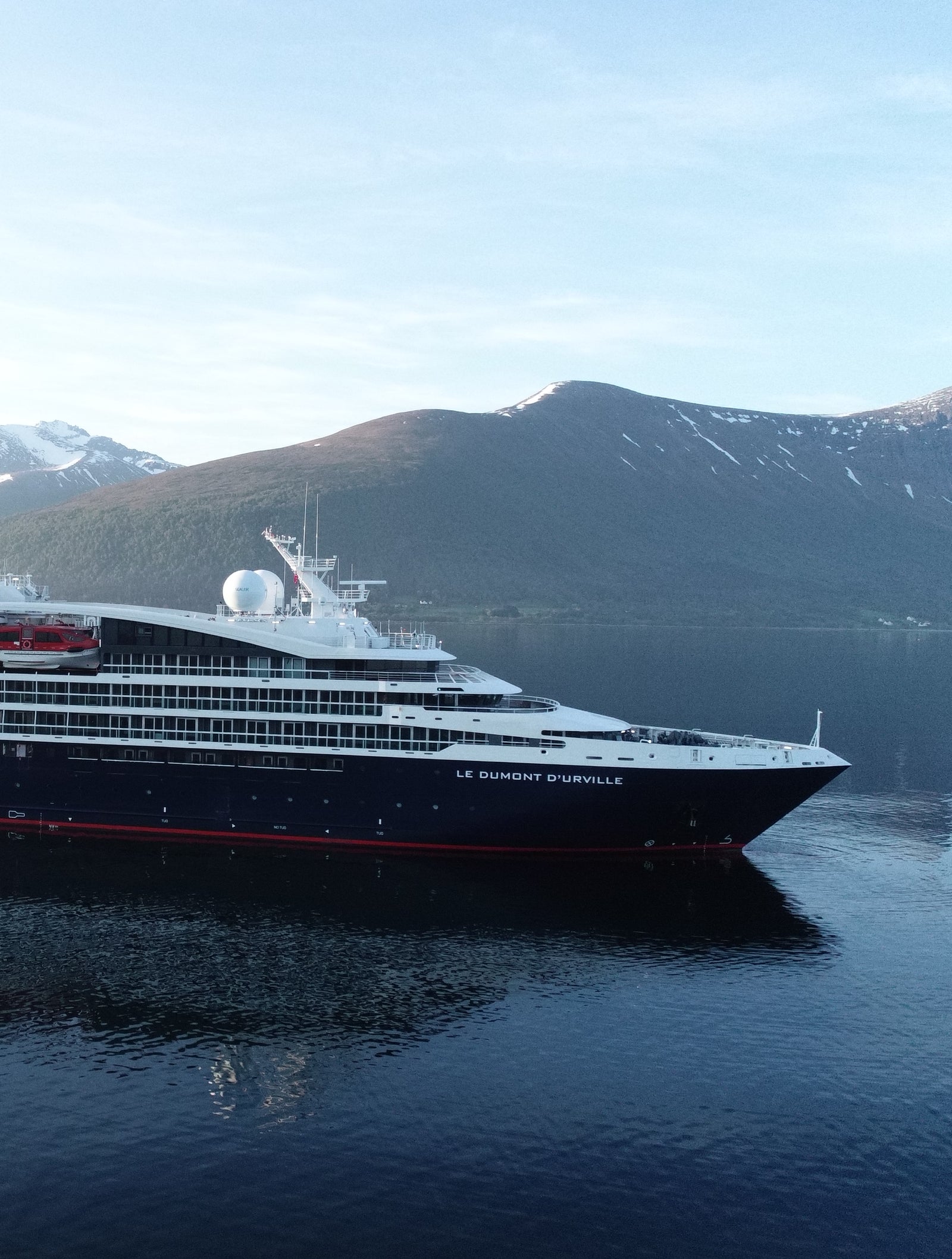 Fincantieri | Gruppo - Business - Prodotti - Navi da Crociera - Le Dumont d'Urville: Vista laterale della nave Le Dumont d'Urville operativa in navigazione all’interno di un fiordo, circondata da montagne innevate e acque calme che riflettono il paesaggio, scritta "Ponant" visibile sullo scafo.