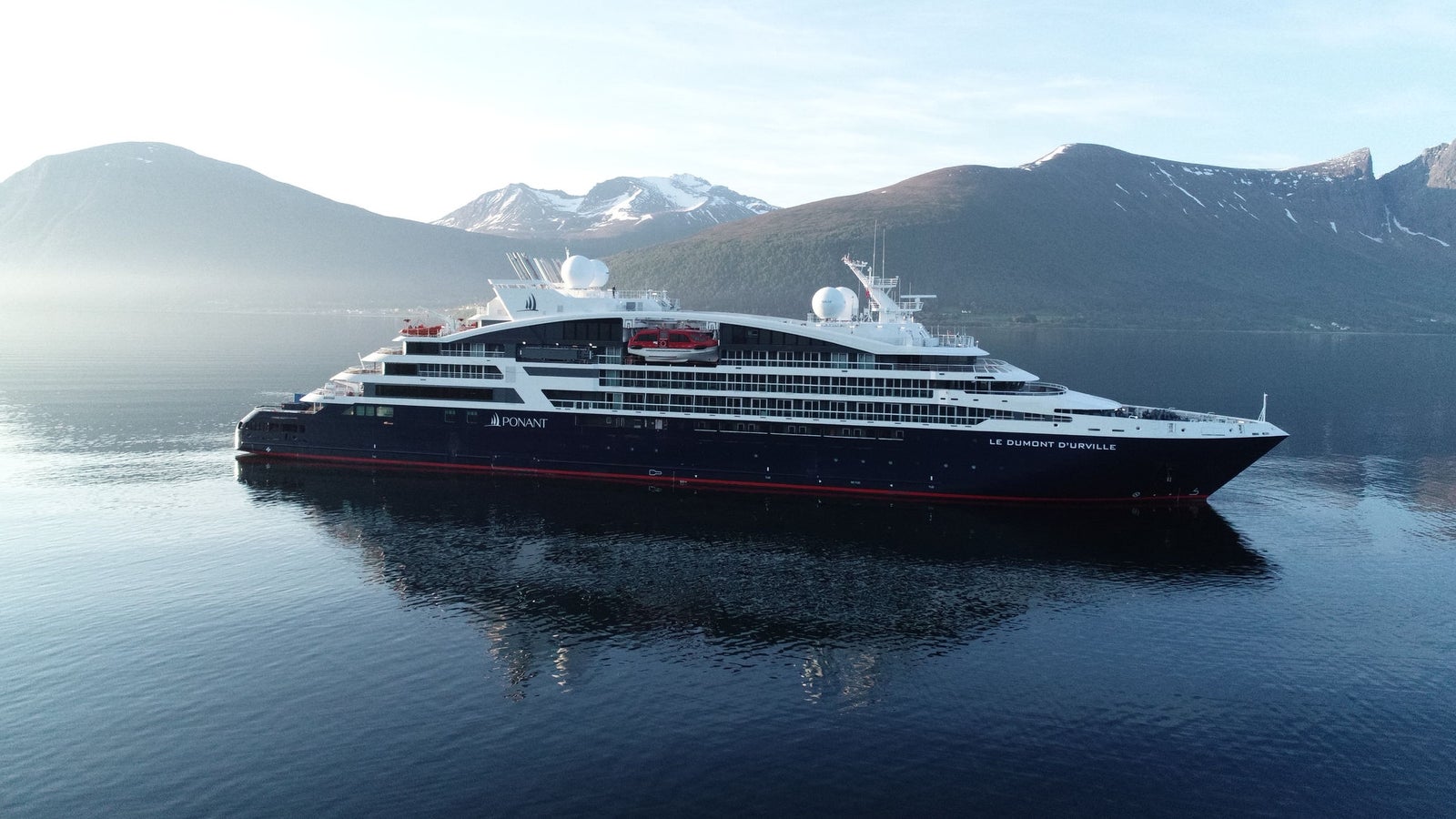 Fincantieri | Gruppo - Business - Prodotti - Navi da Crociera - Le Dumont d'Urville: Vista laterale della nave Le Dumont d'Urville operativa in navigazione all’interno di un fiordo, circondata da montagne innevate e acque calme che riflettono il paesaggio, scritta "Ponant" visibile sullo scafo.