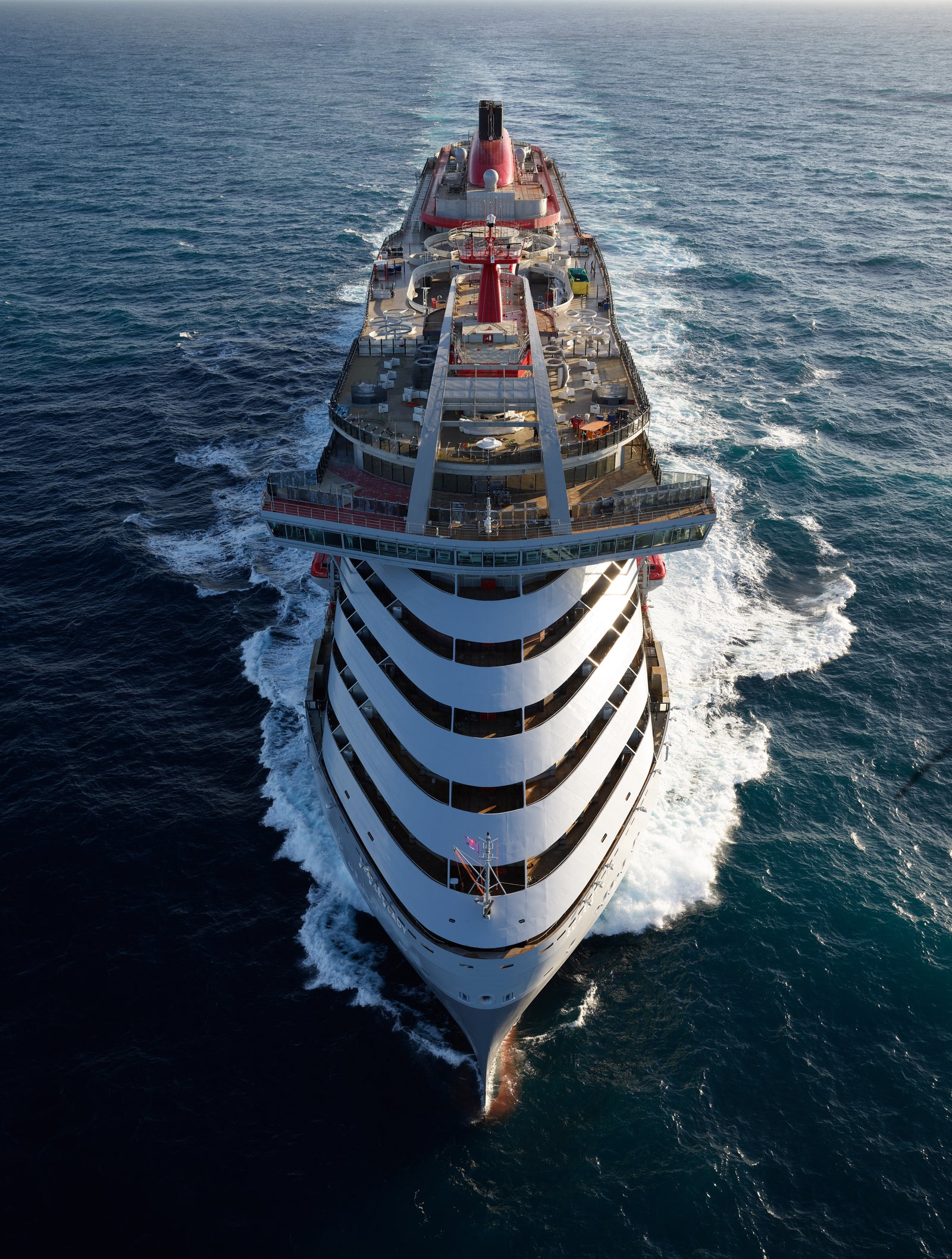 Fincantieri | Group - Business - Products - Cruise Ships - Scarlet Lady: Aerial view of large luxury cruise ship with striped design, spacious decks and relaxation areas, sailing on calm waters under clear sky, serene and adventurous atmosphere.