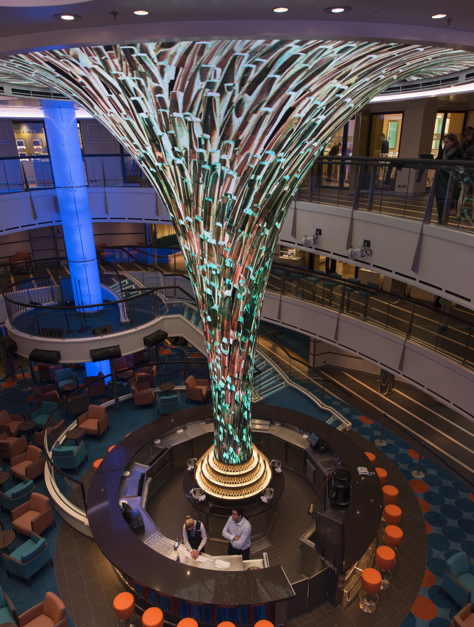 Fincantieri | Group - Business - Products - Cruise Ships - Carnival Vista: Contemporary bar with illuminated central sculpture in spacious multi-level indoor area, seating with vibrant orange stools, decorated floor, lively atmosphere, two staff members visible at the counter.
