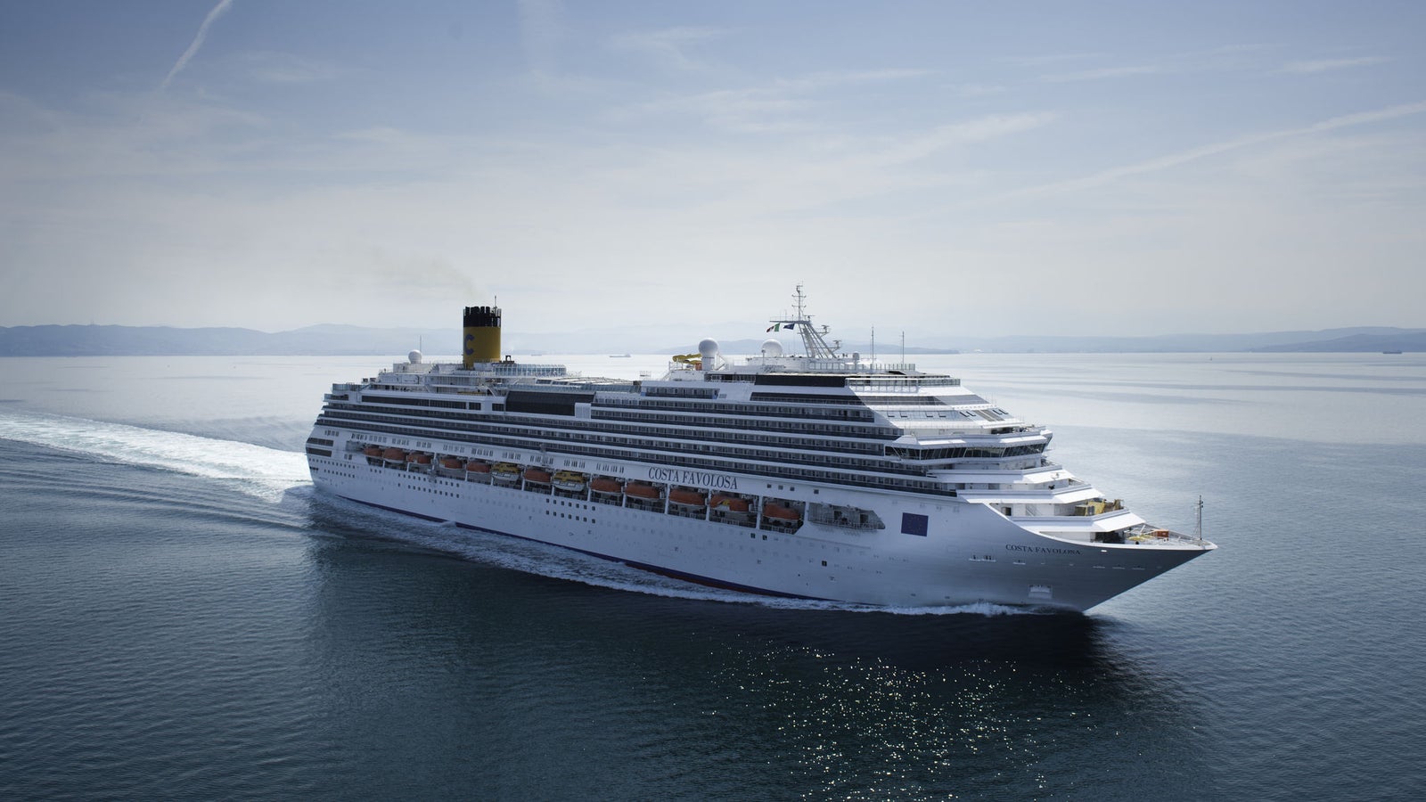 Fincantieri | Group - Business - Products - Cruise Ships - Costa Favolosa: Side view of Costa Favolosa cruise ship in operation, white hull with multiple passenger decks and yellow funnel with black top.