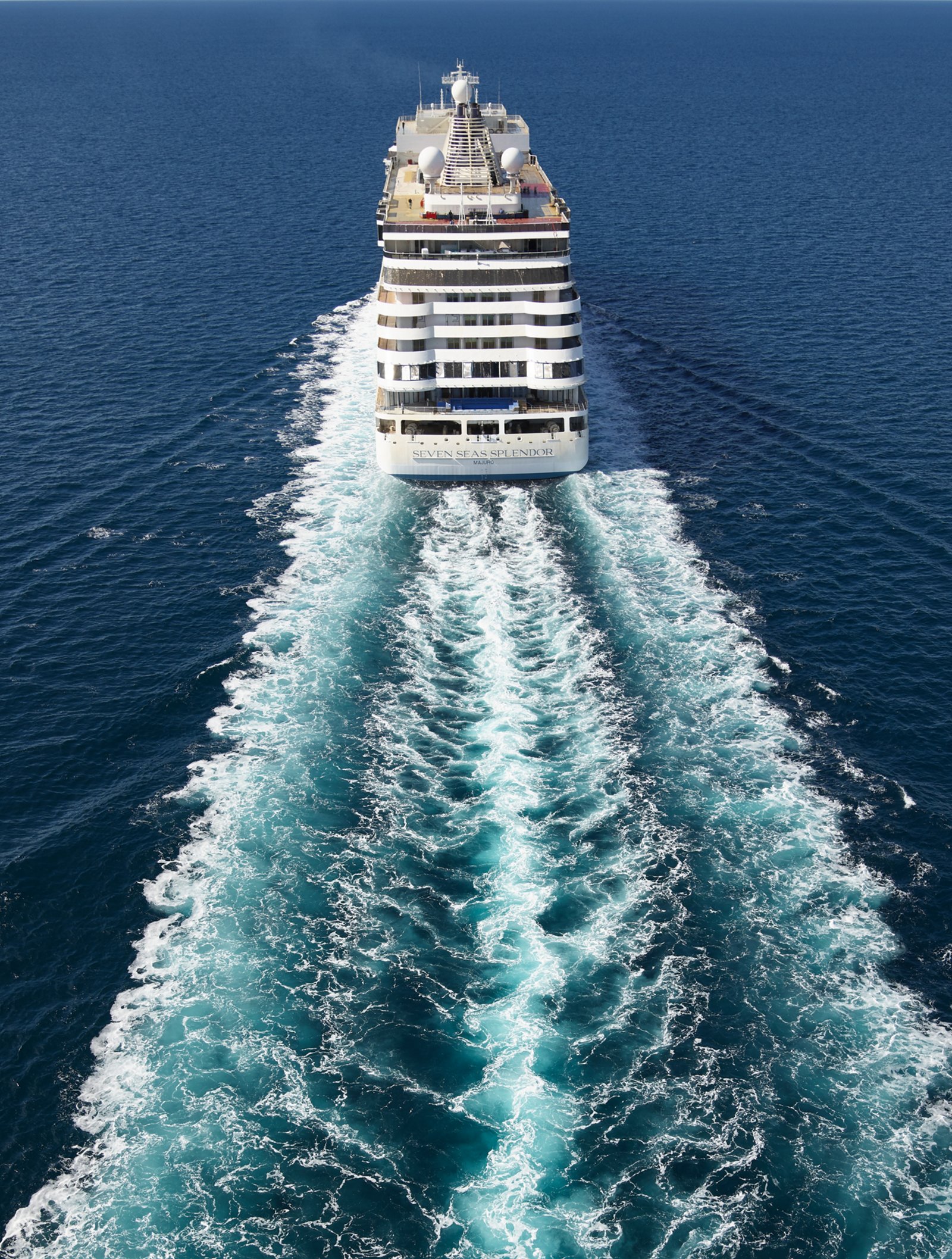 Fincantieri | Group - Business - Products - Cruise Ships - Seven Seas Splendor: Front view of large modern operational cruise ship sailing on calm waters under clear blue sky, visible wake, balconies and multiple decks, atmosphere of exploration and tranquility with wide views of ocean and serene sky.