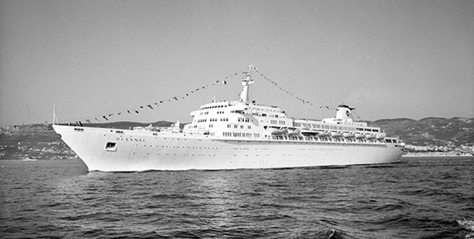Fincantieri | About Us - Historic Ships: Black and white photo of a large cruise ship named Oceanic sailing on calm waters, side view with funnel visible at the stern, mountain chains near the coast in the background.