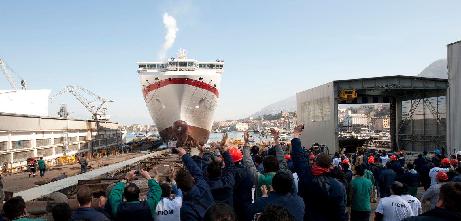 Fincantieri | Group - Business - Products - Ferries - Cruise Europa: Large ship being launched at dockyard, surrounded by crowd of workers cheering; industrial cranes and dock facilities present, clear sky and distant mountains visible; ship’s bow has red and white accents, steam rising from chimney.