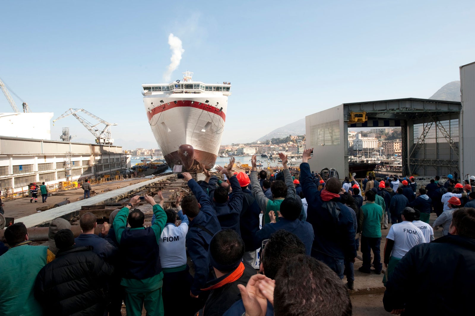 Prodotto - Traghetto Europa 8 Minoan Lines: Vista frontale della nave in fase di varo presso il cantiere navale, folla di operai festanti, gru e strutture industriali, prua con accenti rosso e bianco, vapore dal fumaiolo, sfondo montuoso e cielo sereno.