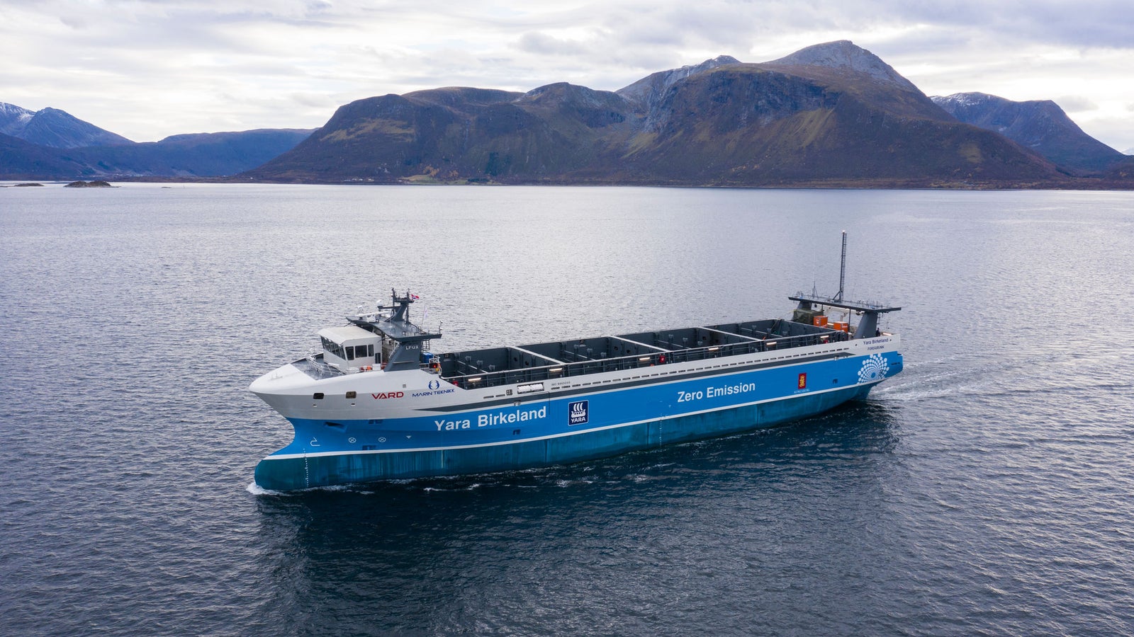 Prodotto - Nave Cargo Yara Birkeland: Vista laterale della nave operativa in navigazione, scritte Yara Birkeland e Zero Emission visibili sullo scafo, sfondo montuoso, design a zero emissioni.