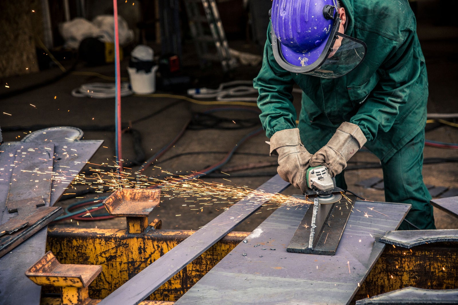 Fincantieri | Business Services - After-Sales Services - Lifecycle Management: A worker wearing protective gear operates an angle grinder on a metal sheet in an industrial workshop. Sparks fly as the tool cuts through the material, emphasizing the intensity of the task. The setting includes heavy-duty equipment and safety measures, highlighting a professional work environment.