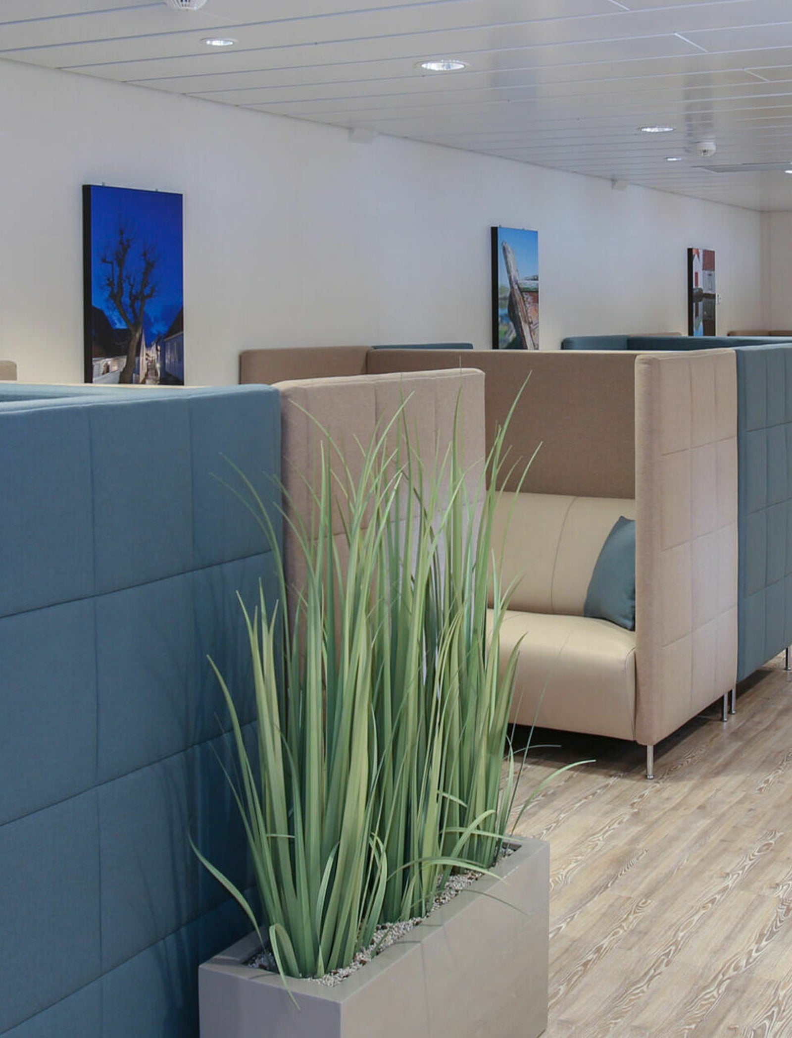 Fincantieri | Gruppo - Società - Controllate e collegate - Vard Interiors: Lounge contemporanea con poltrone box beige e blu per privacy, quadri paesaggistici sulle pareti e vaso decorativo con erba verde alta. Pavimento in legno che aggiunge calore a un design pulito e minimalista.