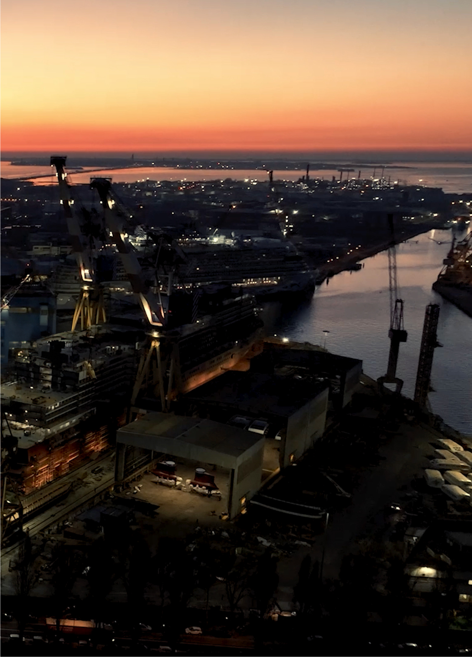 Fincantieri | Group - Sustainability - People and Community - Our People: Large shipyard captured at sunset, big cranes and ships in the foreground, orange light in the background contrasting with dark silhouettes of machinery; calm water channel reflects the fading daylight.