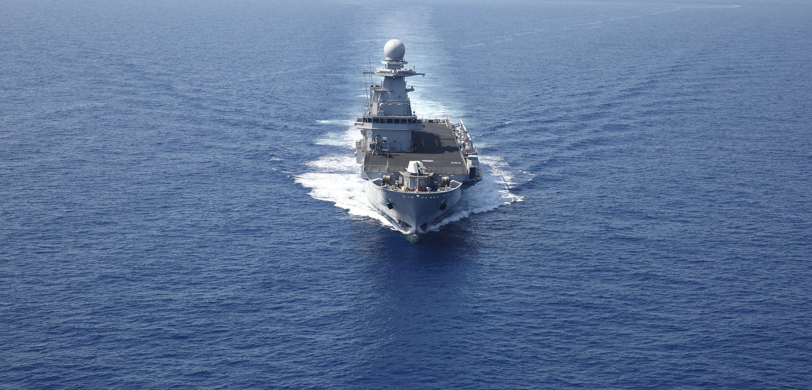Fincantieri | Gruppo - Business - Prodotti - Navi Militari - Portaerei e Navi Anfibi - Kalaat Beni Abbes: Vista frontale della portaerei "Kalaat Beni Abbes" in operazione, in navigazione su acque blu calme, scia bianca visibile, in avvicinamento verso la fotocamera.