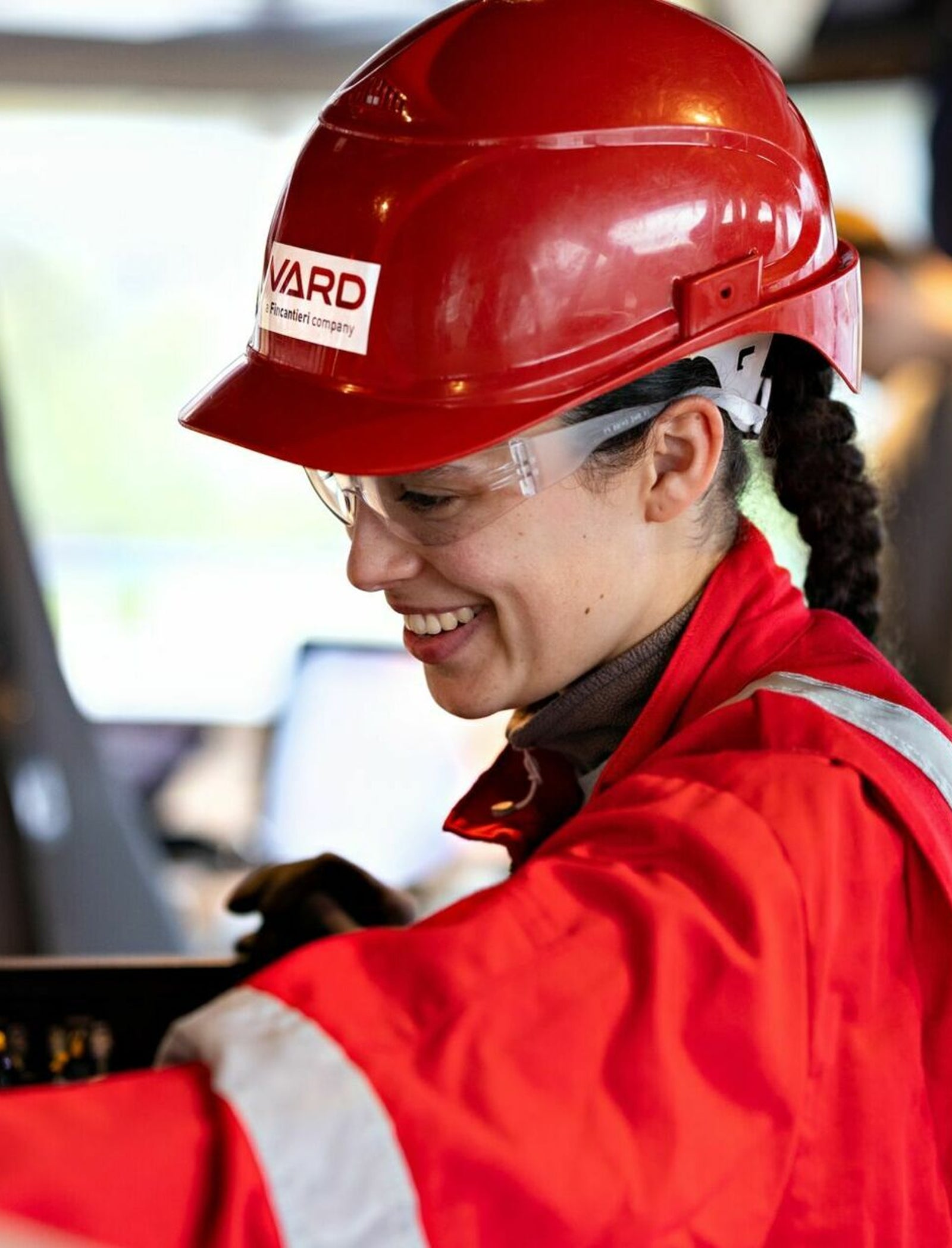 Fincantieri | Group - Subsidiaries and Affiliates - Vard Electro: Person wearing a red helmet and safety uniform working at a control station, ‘VARD’ text clearly visible on helmet; industrial setting with blurred background machinery, focus on task in professional environment.