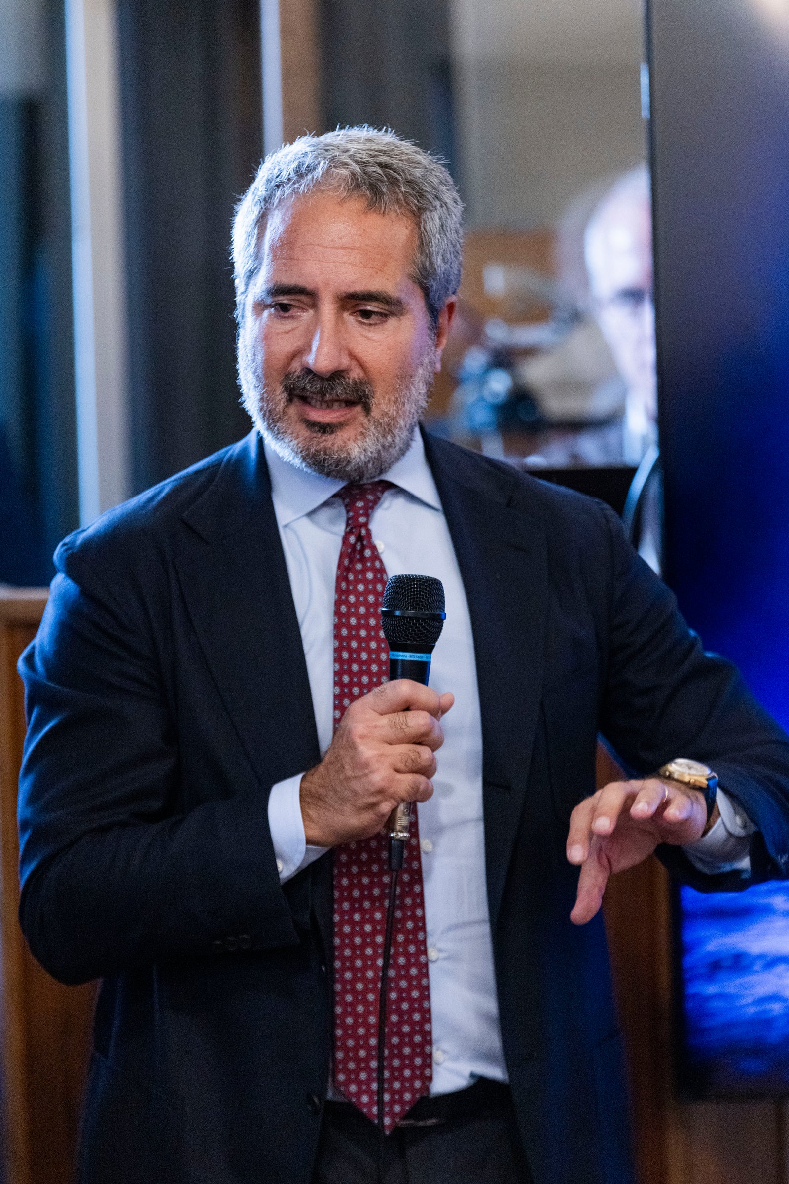 Fincantieri | Newsroom, Deep: Pierroberto Folgiero wearing a formal suit and tie is captured mid-speech at an event. The setting features a blue-lit background, suggesting a corporate or professional environment. The individual is gesturing with their hand, emphasizing their point.