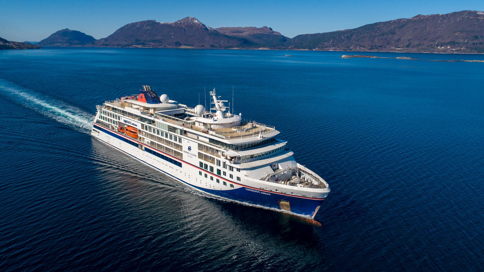Fincantieri | Gruppo - Business - Prodotti - Navi da Crociera - HANSEATIC nature: Nave da crociera moderna operativa che scivola su acque blu calme circondata da paesaggi montuosi, più ponti e scialuppe di salvataggio visibili, cielo limpido e ambiente sereno valorizzano la scena.