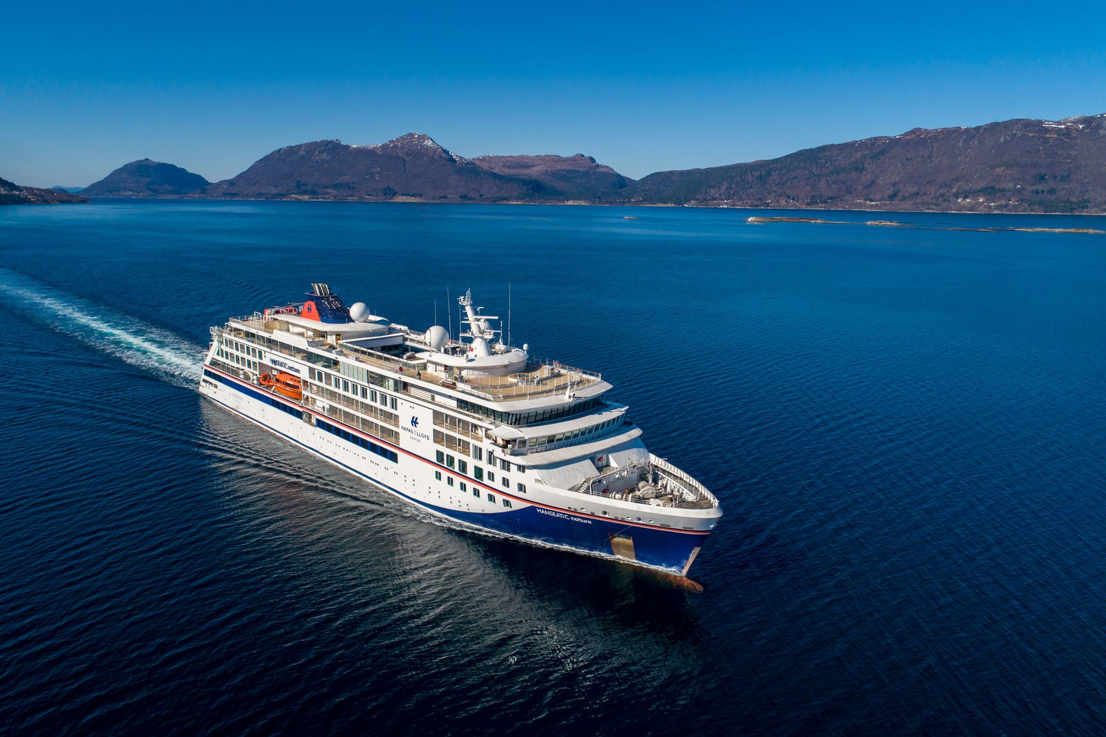 Fincantieri | Gruppo - Business - Prodotti - Navi da Crociera - HANSEATIC nature: Nave da crociera moderna operativa che scivola su acque blu calme circondata da paesaggi montuosi, più ponti e scialuppe di salvataggio visibili, cielo limpido e ambiente sereno valorizzano la scena.