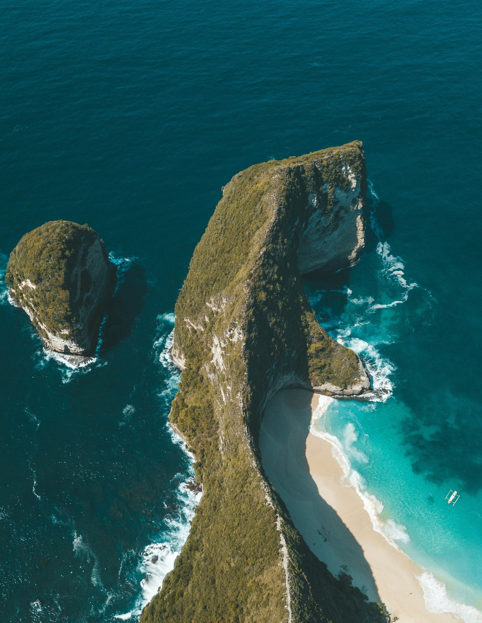 Fincantieri | Sustainability - Energy Transition: Aerial view of a rock formation extending into the turquoise ocean, curved white sandy beach at the base of the cliff.