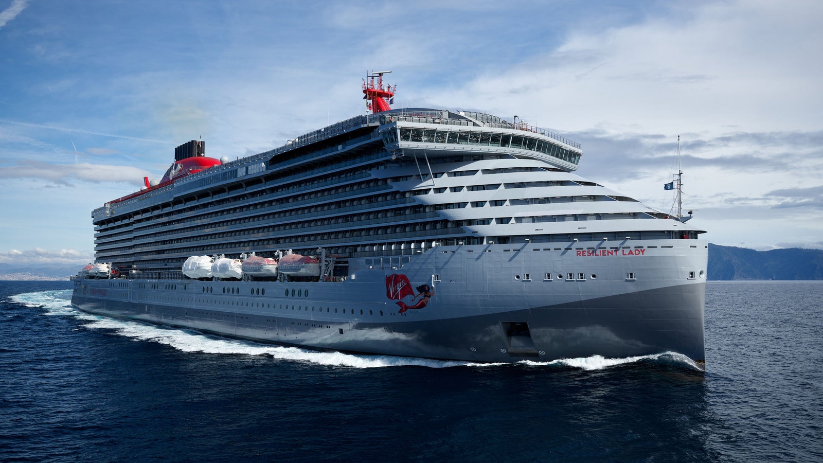 Fincantieri | Group - Business - Products - Cruise Ships - Resilient Lady: Large Virgin Voyages operational cruise ship with modern design and prominent red details, multiple decks, name 'Resilient Lady' visible on hull, sailing in calm waters under partly cloudy sky, maritime elegance.