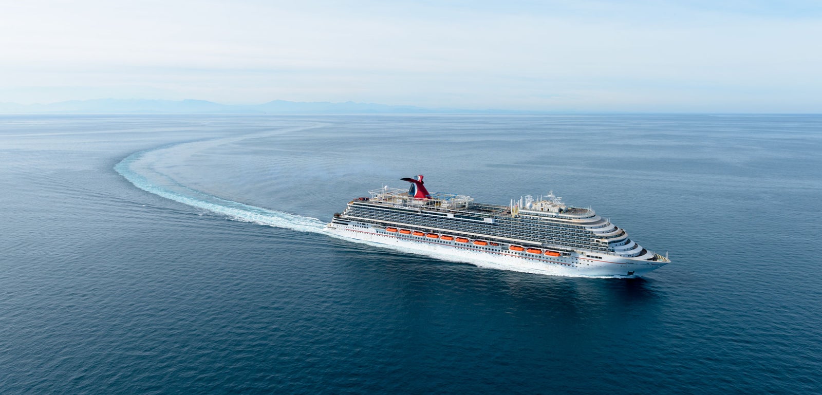 Fincantieri | Group - Business - Products - Cruise Ships - Carnival Horizon: Massive operational cruise ship moving through calm open waters under clear sky, multiple decks with vibrant red and white accents contrasting with blue sea, curved visible wake highlights movement and ship size.