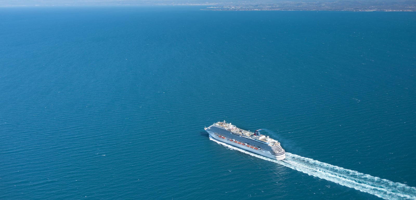 Fincantieri | Group - Business - Products - Cruise Ships - Carnival Magic: Large cruise ship in operation navigating vast blue ocean under clear sky, visible wake behind showcases speed and direction, faint horizon adds depth to serene maritime scene.