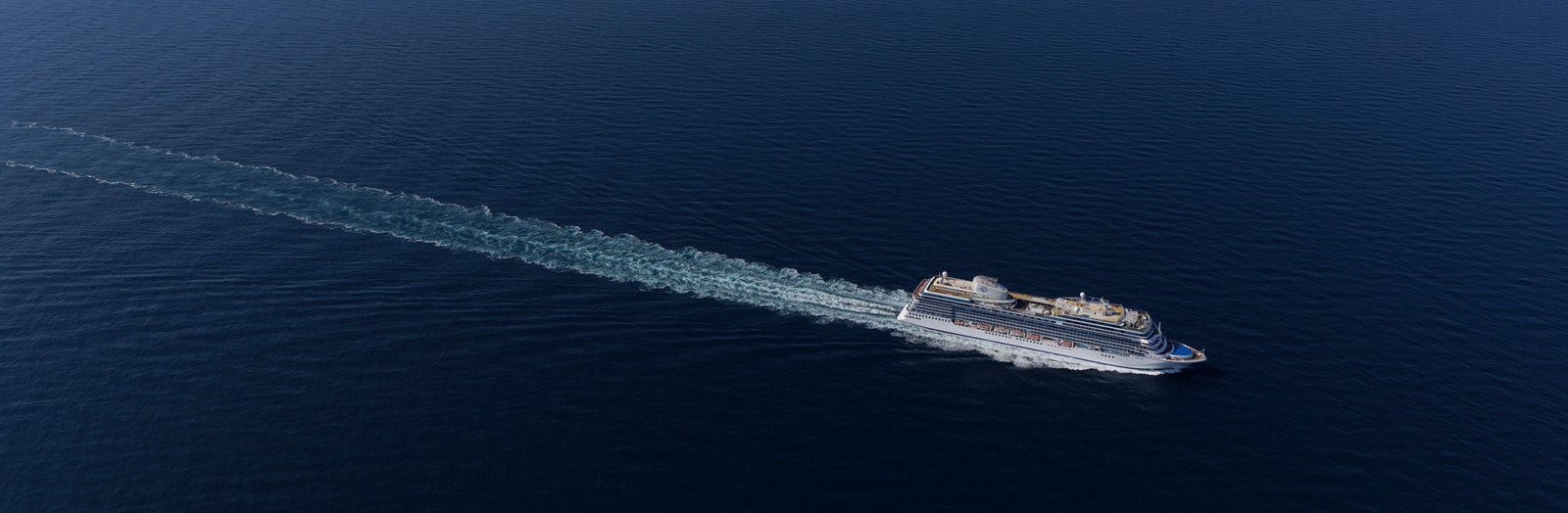 Fincantieri | About Us - Strategic Vision: Aerial view of a large operational cruise ship sailing on the open ocean, visible wake in the water, calm marine environment, and clear blue sky.