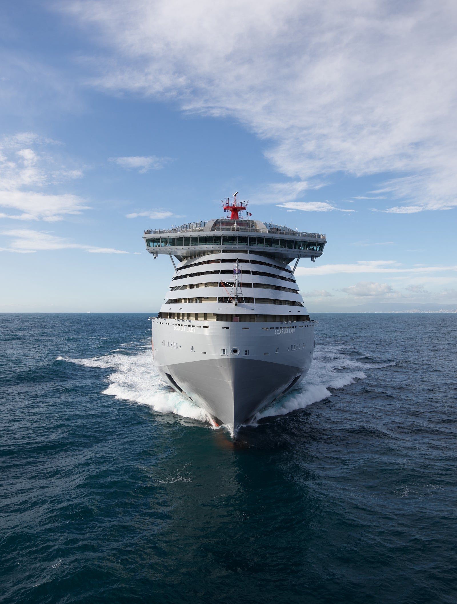 Fincantieri | Group - Business - Products - Cruise Ships - Scarlet Lady: View of modern bow of Scarlet Lady cruise ship in operation, elegant and distinctive design with red mermaid motif and smoked glass, sailing on calm water under partly cloudy sky, atmosphere of adventure in open sea.