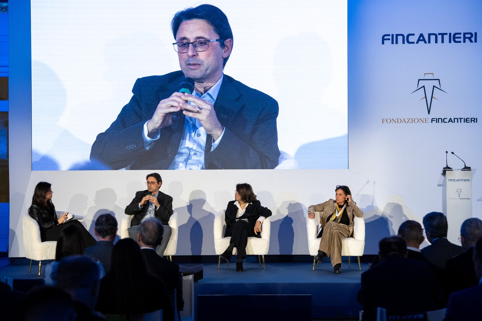Newsroom - Comunicati stampa - 2025 - Gruppo Fincantieri: Un anno di Fondazione Fincantieri: storia, innovazione e impatto sociale: Panel di ospiti e relatori seduti sul palco durante l’evento Fondazione Fincantieri, con maxi schermo, loghi istituzionali e pubblico in sala.