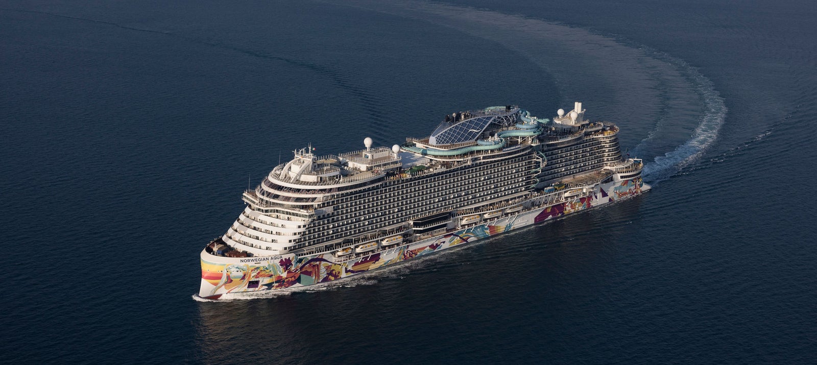 Fincantieri | Group - Innovation - Naval Ecosystem of the Future: Large operational cruise ship sailing on calm waters under an overcast sky, colorful decorations on the side, multiple decks with visible pools and relaxation areas, distinct wake highlighting the ship’s size and power.