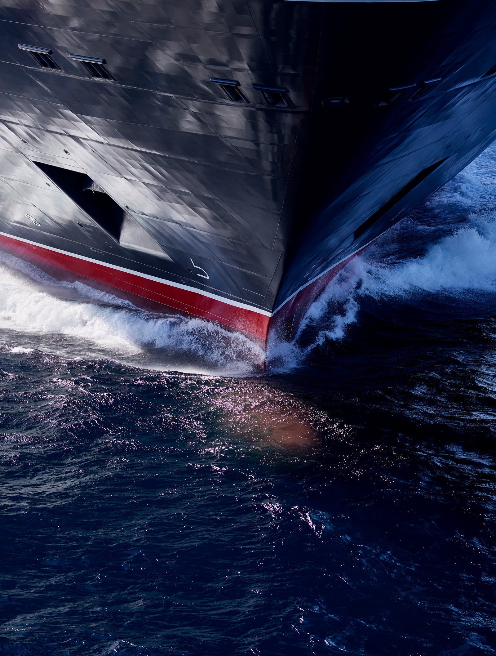 Fincantieri | Group - Business - Products - Cruise Ships - Queen Anne: Close-up of cruise ship bow in operation, cutting through water with spray, dark hull with red detail along side, open sea.