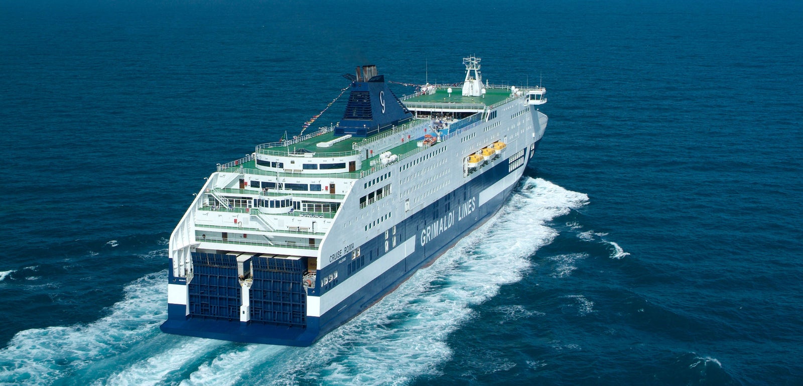 Fincantieri | Group - Business - Products - Ferries - Cruise Roma: Large "Grimaldi Lines" ferry in operation, multiple decks, visible lifeboats, prominent ship name and branding, open sea, distant coastline, clear blue sky.