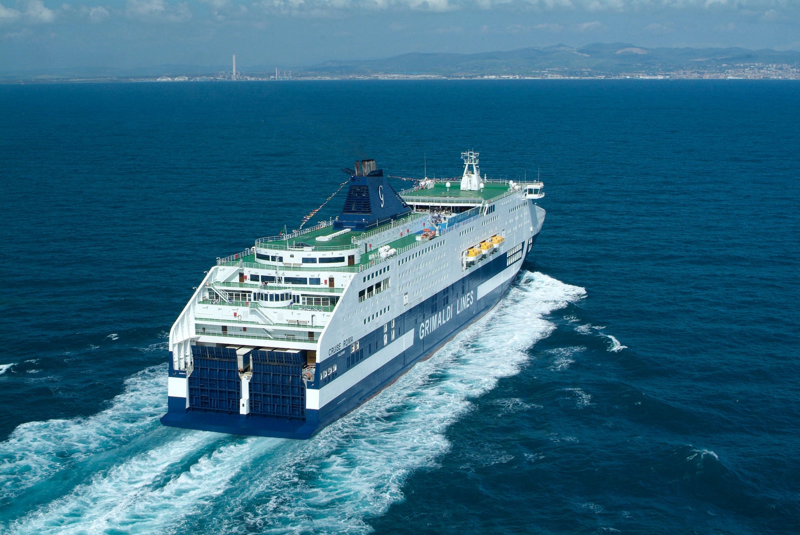 Fincantieri | Group - Business - Products - Ferries - Cruise Roma: Large "Grimaldi Lines" ferry in operation, multiple decks, visible lifeboats, prominent ship name and branding, open sea, distant coastline, clear blue sky.