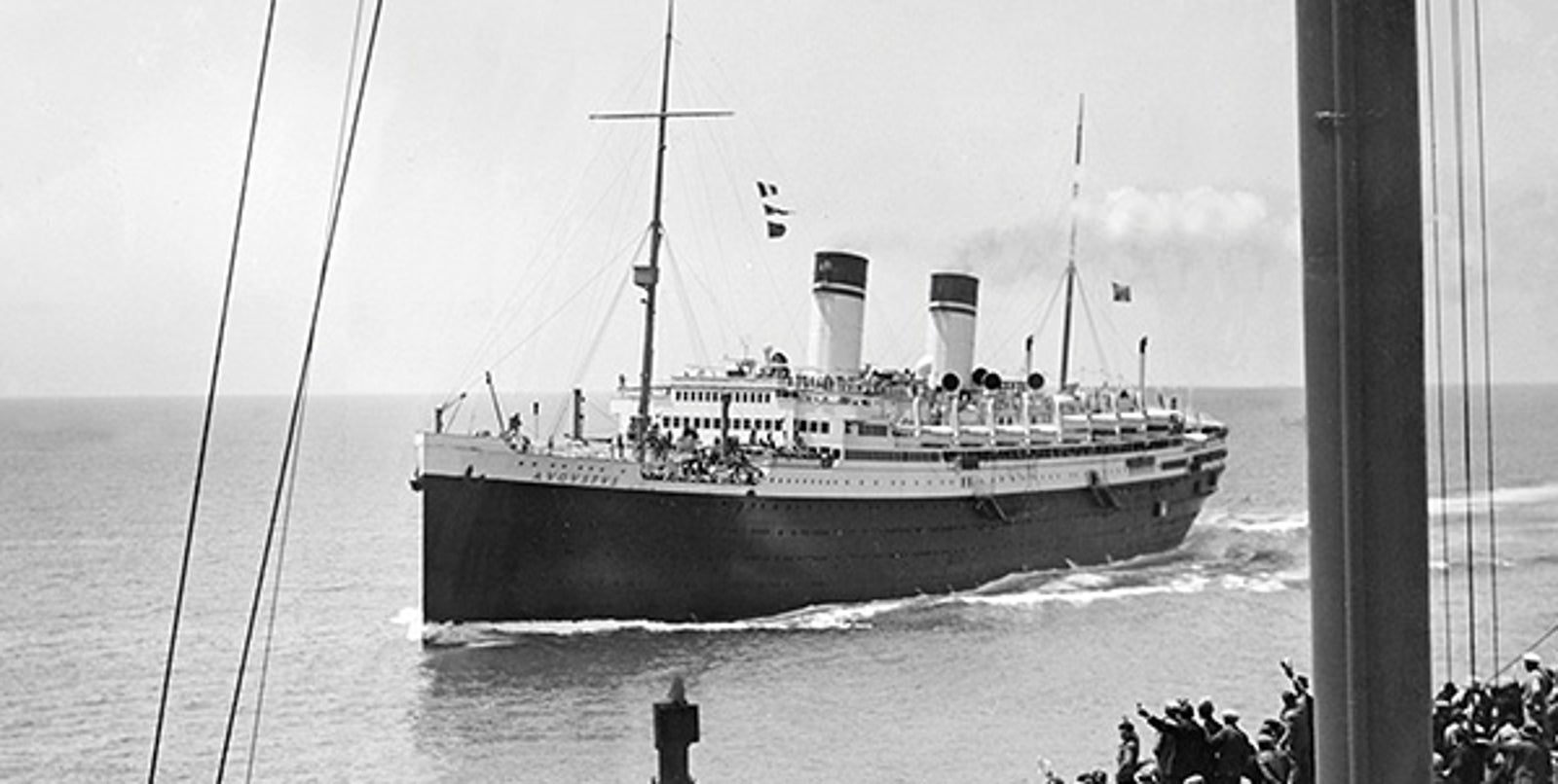 Fincantieri | About Us - Historic Ships: Large historic ocean liner with two funnels sailing on calm waters, visible flags and crowd in the foreground, black and white photo highlighting the classic design and maritime setting.