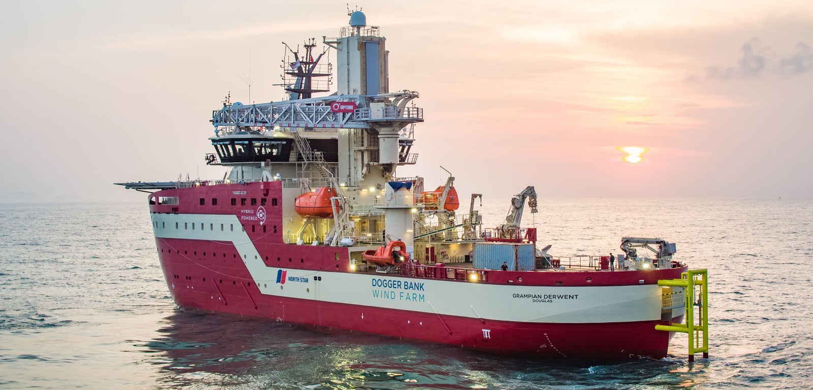 Prodotto - Offshore Support Vessel Derwent: Vista laterale della nave operativa con scafo rosso e bianco, scritta Dogger Bank Wind Farm visibile, gru e piattaforme per operazioni energetiche, navigazione al tramonto, atmosfera tranquilla.