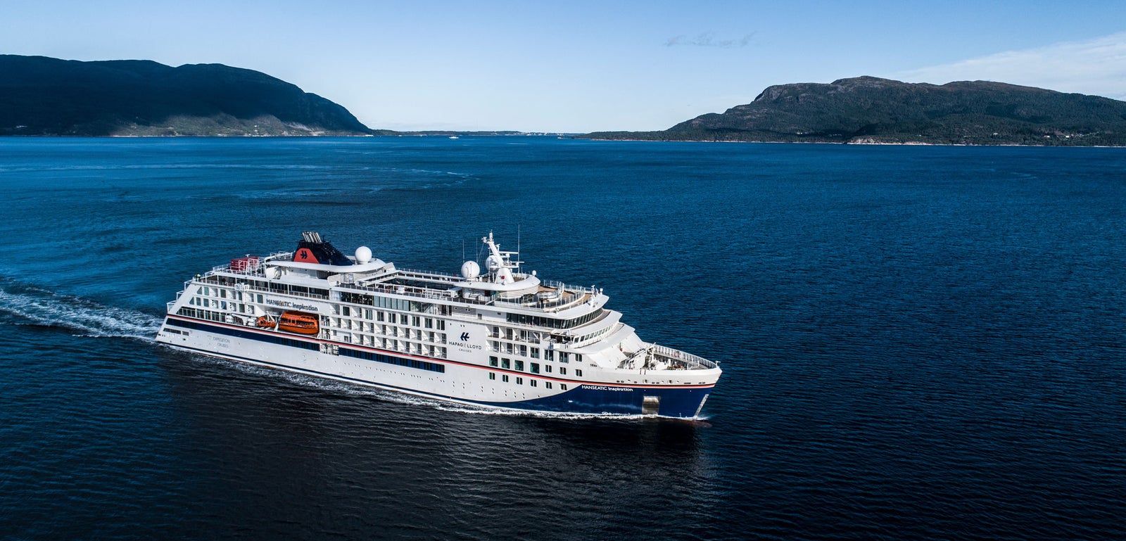  Fincantieri | Gruppo - Business - Prodotti - Navi da Crociera - Hanseatic Inspiration: Grande nave di lusso operativa in navigazione su acque calme blu, molteplici ponti e scialuppe visibili, sfondo con costa montuosa, scena che trasmette eleganza e bellezza naturale.