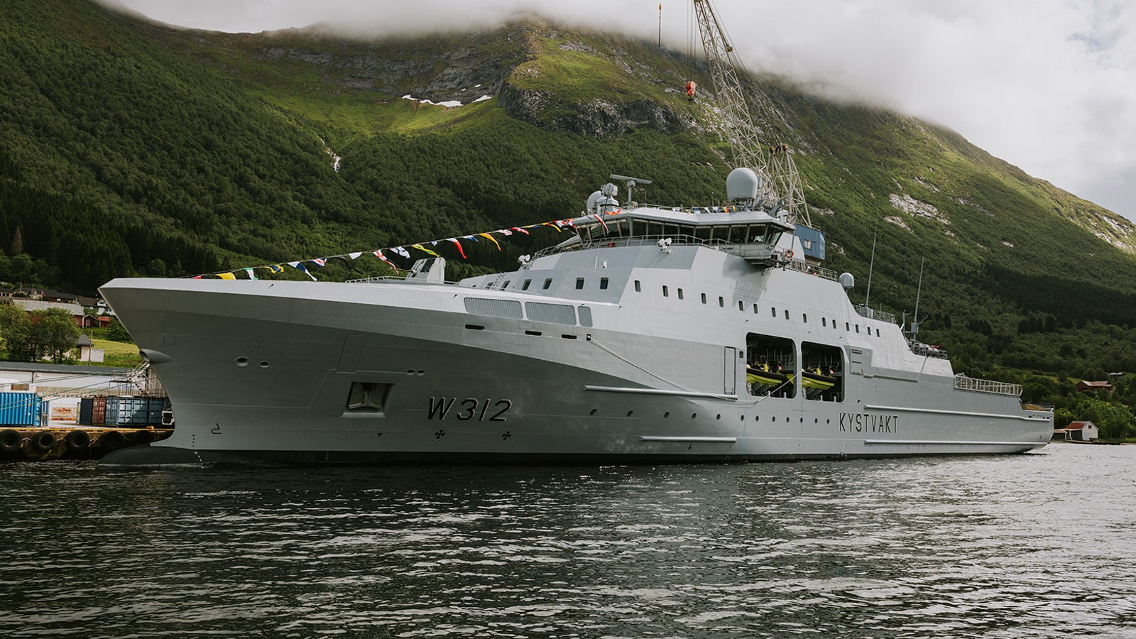 Fincantieri | Group - Business - Products - Defence Vessels - Patrol Vessels - KV Hopen: Side view of vessel docked in port, number W312 visible, modern design, colorful flags along structure, green mountains in background.