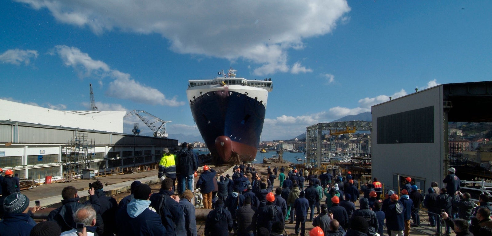Fincantieri | Group - Business - Products - Ferries - Cruise Barcellona: Massive ship being launched at bustling dockyard, surrounded by crowd of workers and spectators; industrial structures, cranes, and clear blue skies; workers in safety gear including helmets and jackets, emphasizing professional maritime environment.