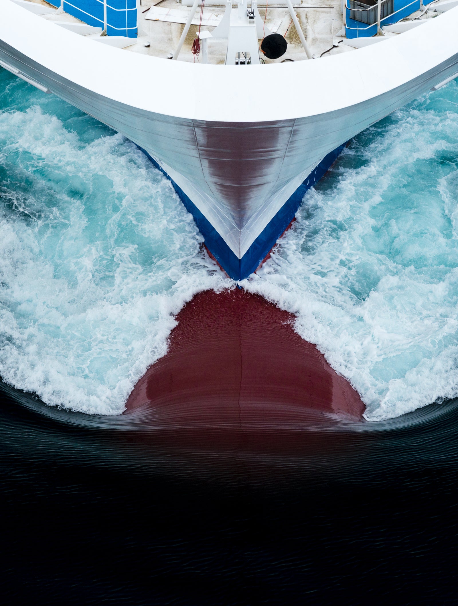  Fincantieri | Group - Business - Products - Cruise Ships - Costa Diadema: Aerial view of Costa Diadema cruise ship bow in operation slicing through deep blue ocean waters, dynamic white foam patterns, partially visible red underside of hull, bold visual contrast.