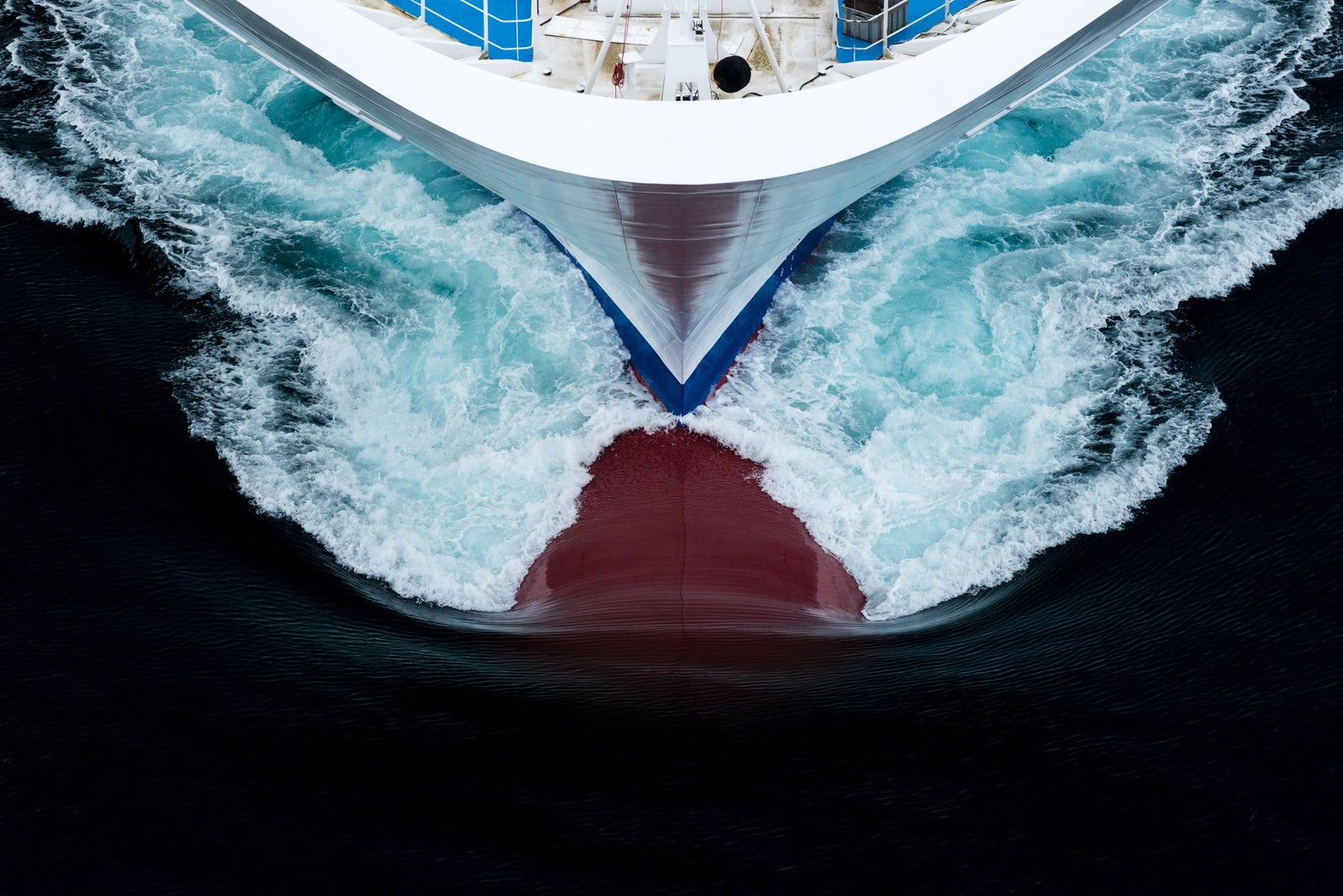  Fincantieri | Group - Business - Products - Cruise Ships - Costa Diadema: Aerial view of Costa Diadema cruise ship bow in operation slicing through deep blue ocean waters, dynamic white foam patterns, partially visible red underside of hull, bold visual contrast.